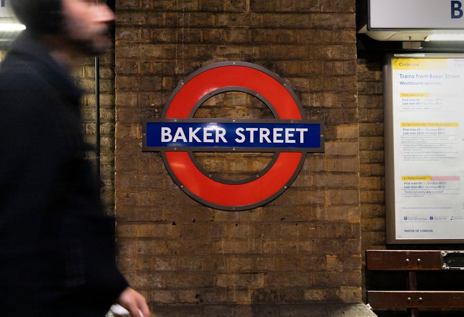 A person with dark hair, wearing a black jacket, is walking past a brick wall inside a station or underground platform. Prominently mounted on the wall is a round London Underground sign with a red ou