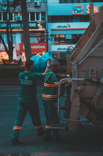 Two waste management workers are operating a large, industrial rubbish collection vehicle on a city street with commercial storefronts and illuminated signage in the background. The worker on the left